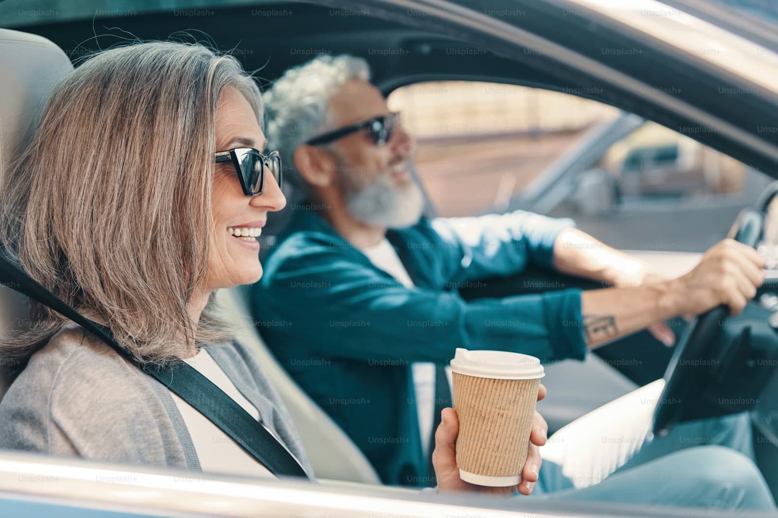 Older couple on a daytime drive, the passenger holding coffee while a gray-bearded man drives